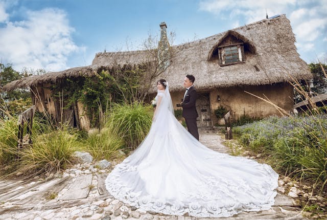 A bride and groom pose in front of a thatched house, showcasing a navy blue dress accessorized for a wedding